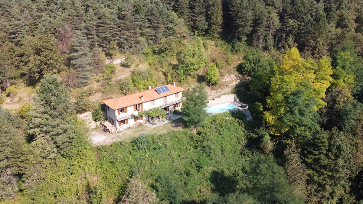 The residence is viewed from above, nestled among lush greenery and trees. The two-story structure features a warm red tile roof and solar panels, with a swimming pool located nearby. The serene natural setting offers a private and tranquil atmosphere.