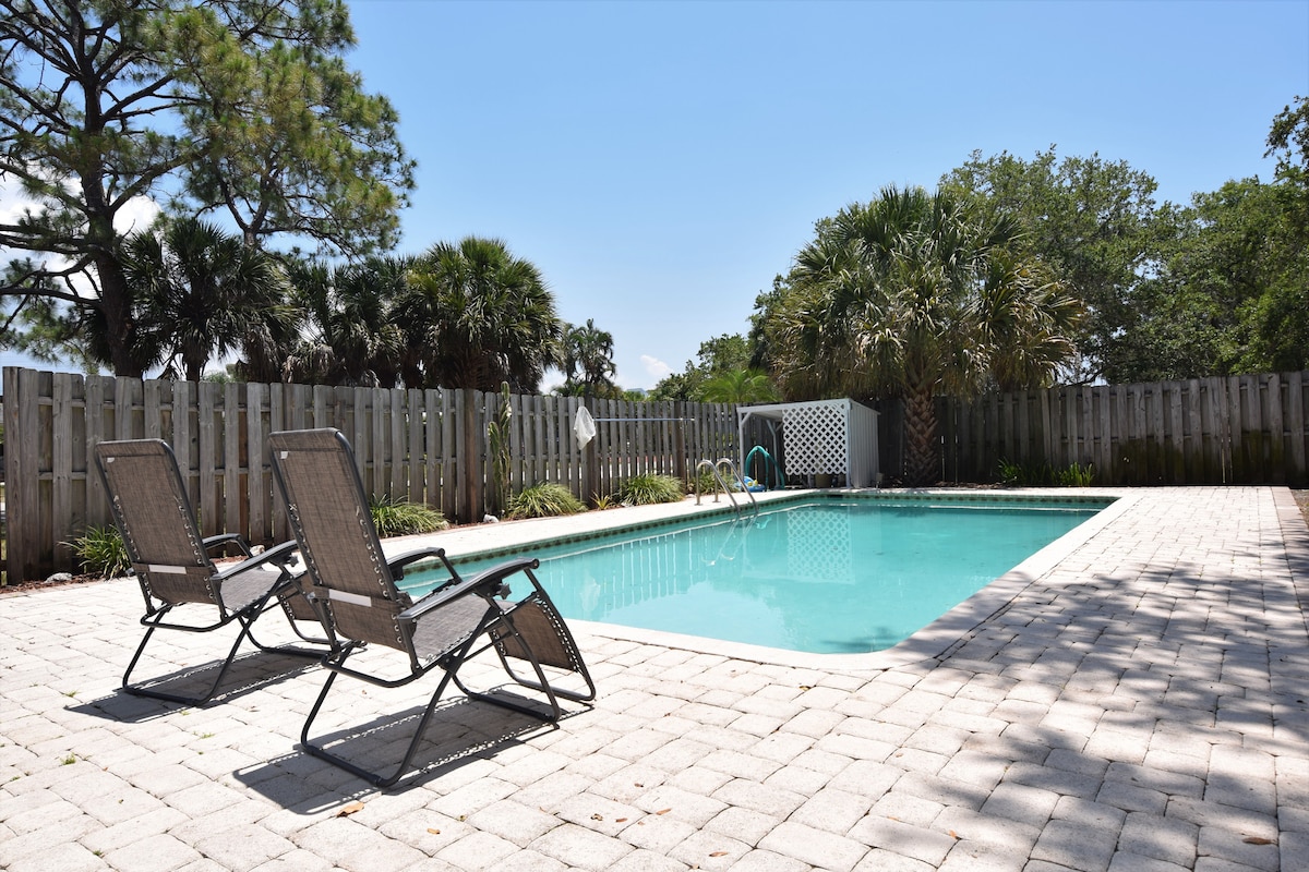 A private fenced pool area is featured, surrounded by greenery and palm trees. Two lounge chairs are positioned beside the clear, inviting water of the pool, which reflects the bright blue sky overhead. The paving stones provide a spacious area for relaxation.