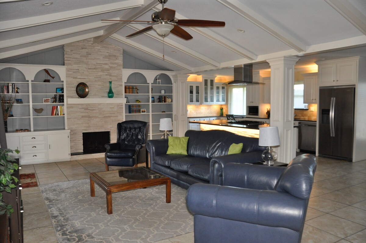 A spacious living area features a comfortable leather seating arrangement, complemented by a coffee table. Built-in shelves display decorative items. A neutral stone fireplace serves as a focal point, while soft lighting highlights the adjacent kitchen area with modern appliances.