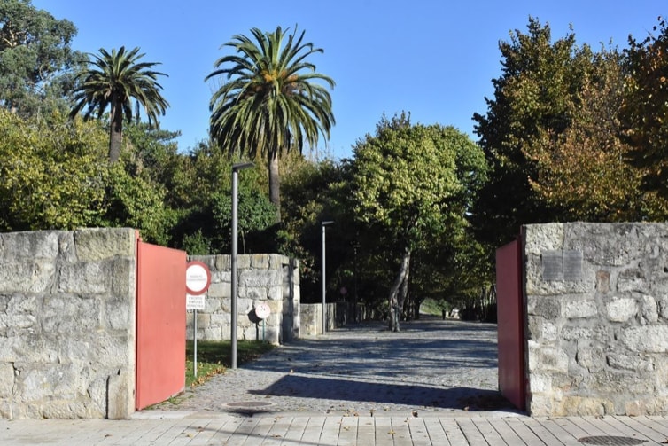 An entrance is framed by tall palm trees and lush greenery, marked by large stone walls. The gates, painted in red, stand open, inviting entry. A clear blue sky provides a bright backdrop, and the path features a mix of cobblestones and paved squares.