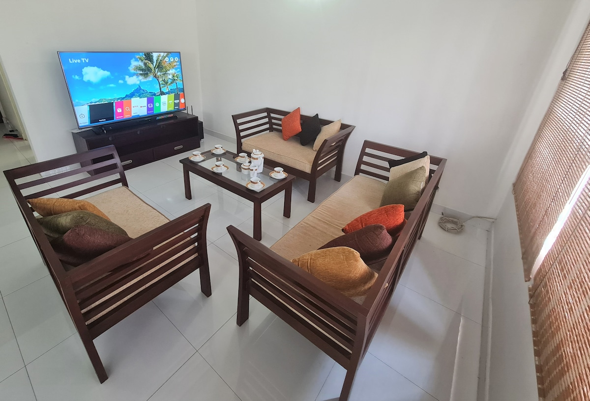 A cozy living area is furnished with four wooden seating arranged around a central coffee table. Decorative cushions in various colors add comfort. A large television is mounted on the wall, providing entertainment options, while natural light streams in through nearby window treatments.