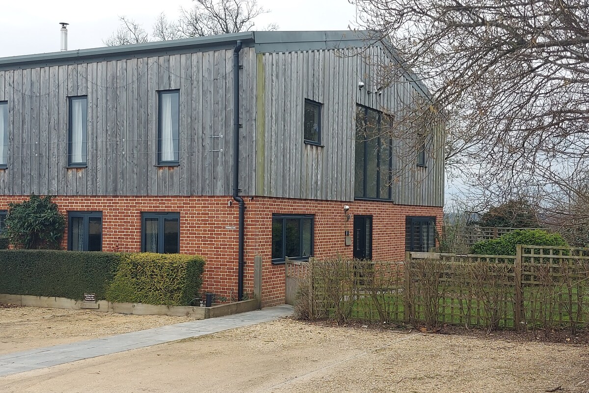 The exterior of a modern barn conversion is showcased, featuring a mix of wooden cladding and red brick. Large windows offer glimpses of the interior, while a neatly maintained garden and hedges surround the entrance. A gravel driveway leads up to the building.