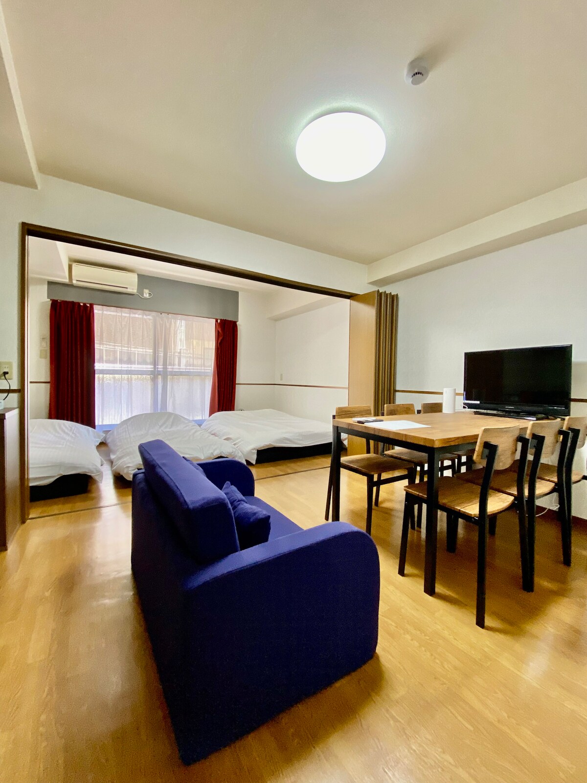 A spacious living area features a sofa and a dining table with chairs. Two beds are arranged in the background. Natural light enters through a large window adorned with curtains. A television can be seen on a side table, creating a functional and comfortable environment.