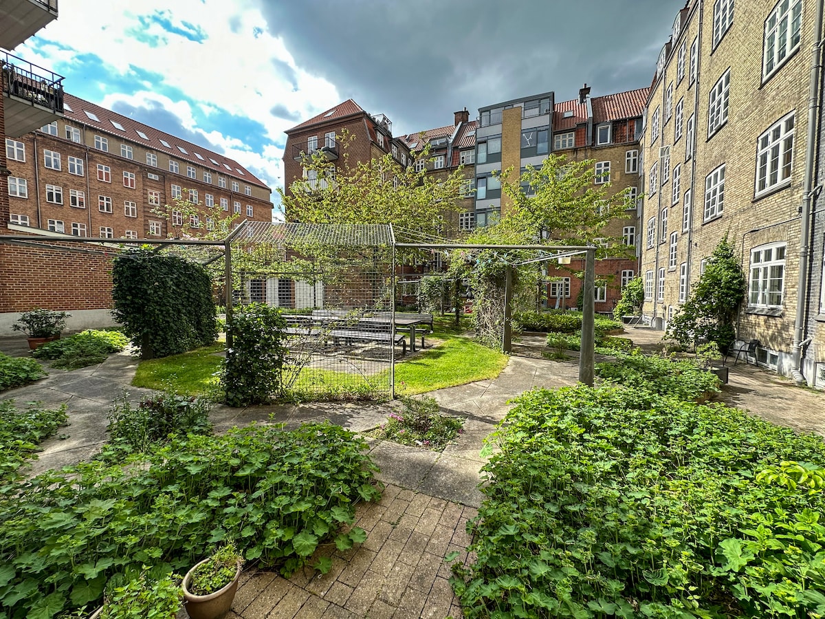 A shared garden area is surrounded by brick buildings, featuring patches of grass and shrubbery. A wooden seating area with tables is situated under a metal pergola. The space is designed for relaxation amidst greenery, providing a quiet outdoor retreat.
