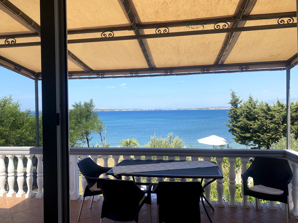 A spacious terrace is visible, featuring a table and several chairs. The expansive view reveals a serene sea landscape, where the water reflects the clear blue sky. Lush greenery frames the scene, creating a calming atmosphere for relaxation.
