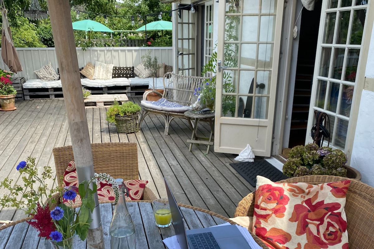 An inviting terrace showcases a wooden deck adorned with potted plants and comfortable seating. A round table with floral arrangements is placed in the foreground, while lounge areas with cushions and umbrellas are visible in the background, creating a relaxing outdoor space.