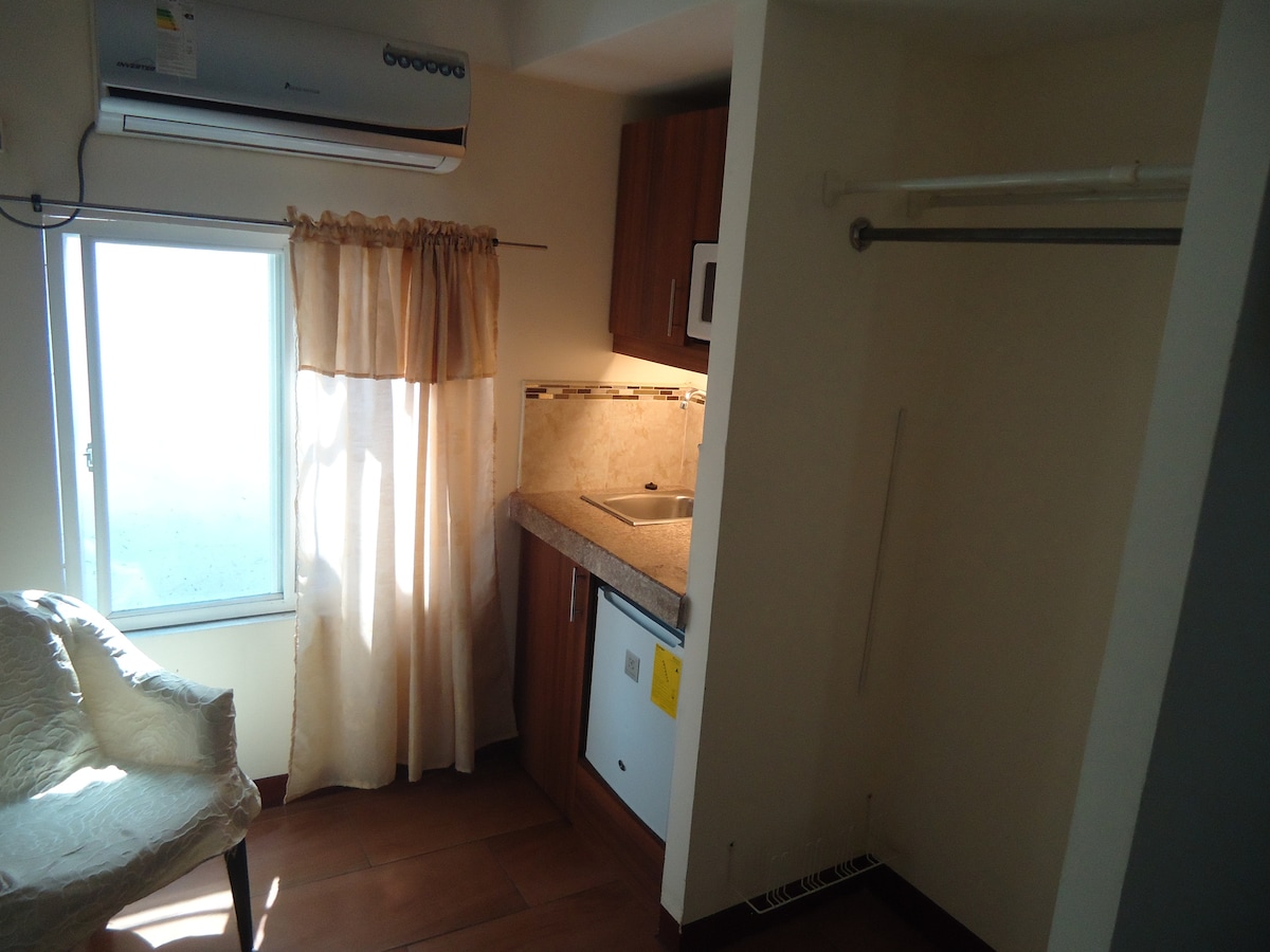 A compact kitchenette features a countertop with a sink and cabinets above. A small refrigerator is positioned below. Natural light filters in through a window adorned with light curtains, complemented by a cozy chair nearby to create an inviting space.