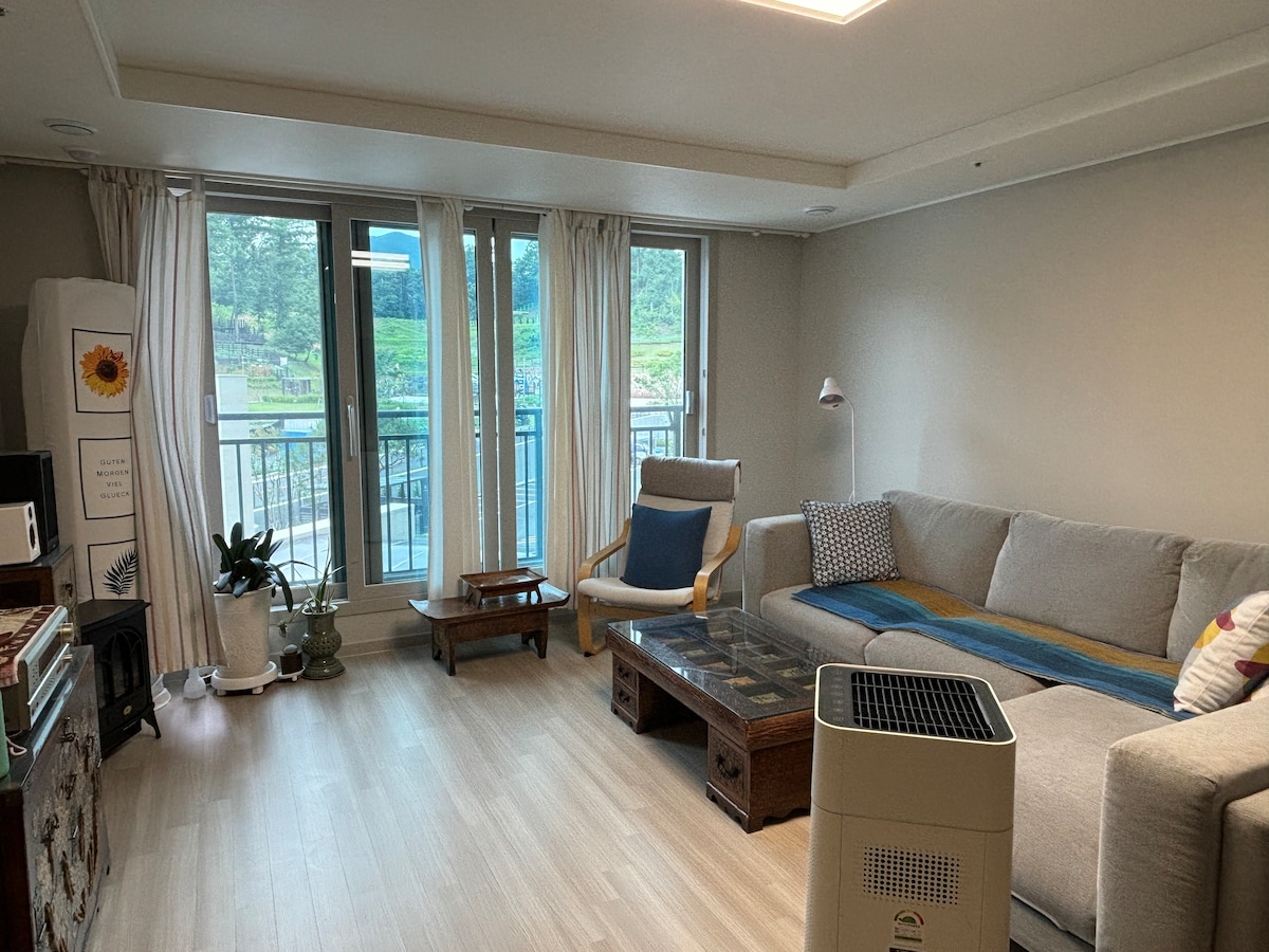 A bright living area features a spacious sofa and a coffee table positioned near large windows. Light curtains frame the view, while a small indoor plant and a lamp provide added comfort. An air purifier sits discreetly on the floor, enhancing the overall ambiance.