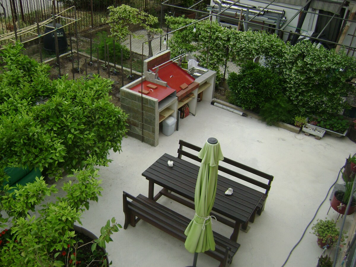 A spacious garden area is visible, featuring a wooden table with benches beneath a green umbrella. A barbeque setup is positioned nearby, along with various plants and shrubs that define the outdoor space, creating a serene environment.