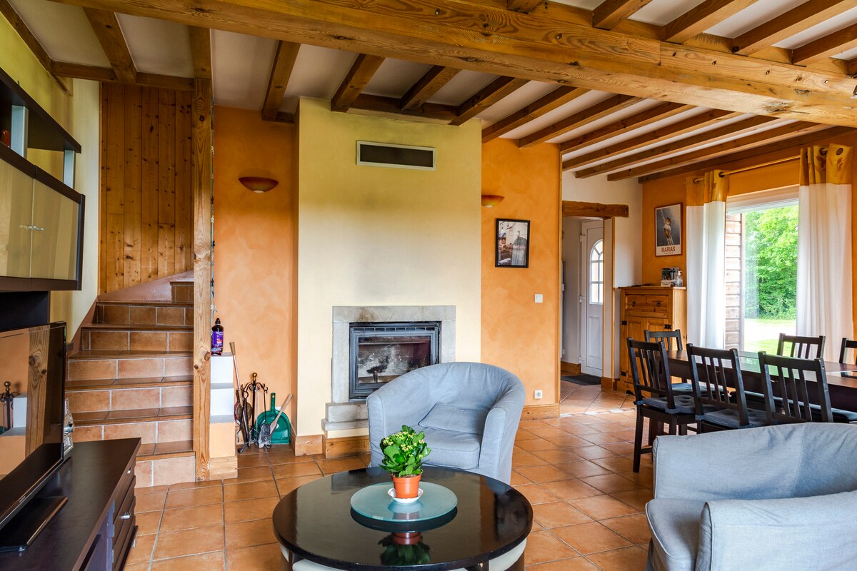 The living area features a cozy seating arrangement with two armchairs and a round table at its center. A fireplace is set against a softly painted wall, while a dining table with chairs is positioned nearby. Large glass doors open to the garden, enhancing natural light.