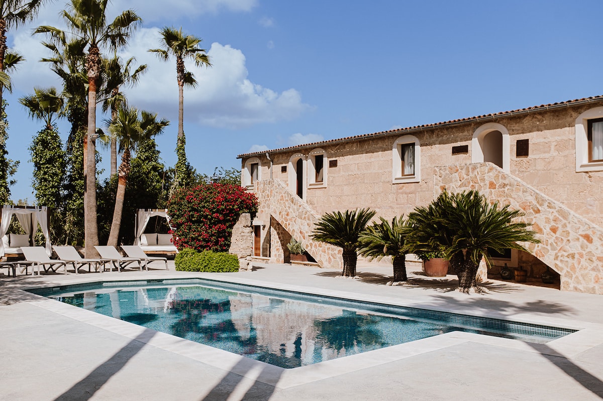 The outdoor area features a rectangular chlorinated pool surrounded by paved surfaces and lounge chairs. Lush palm trees and vibrant foliage provide a tropical ambiance. The two-story building has a natural stone facade with multiple windows, and outdoor staircases lead to the upper level.