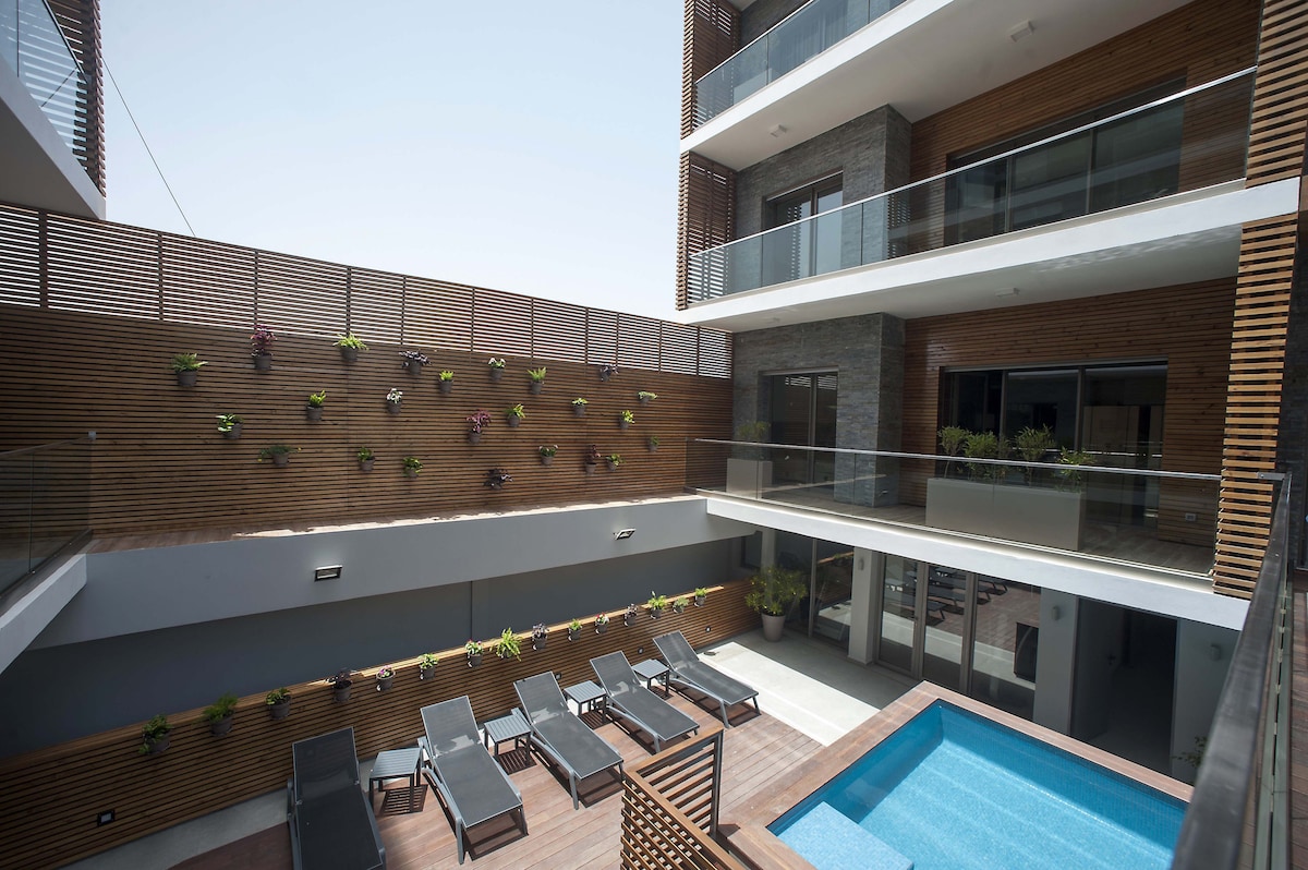 An inviting courtyard is revealed, featuring a small pool surrounded by several lounge chairs. Vertical planters display greenery against wooden paneling, adding a touch of nature. The modern building structure provides multiple stories with balconies overlooking the area, enhancing light and openness.