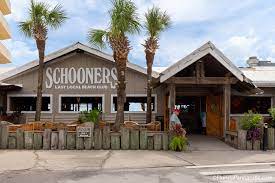 The exterior of Schooners restaurant features a rustic wooden structure with a peaked roof. Lush palm trees frame the entrance, which is adorned with vibrant flowers and greenery. The establishment showcases a welcoming sign and offers outdoor seating options.