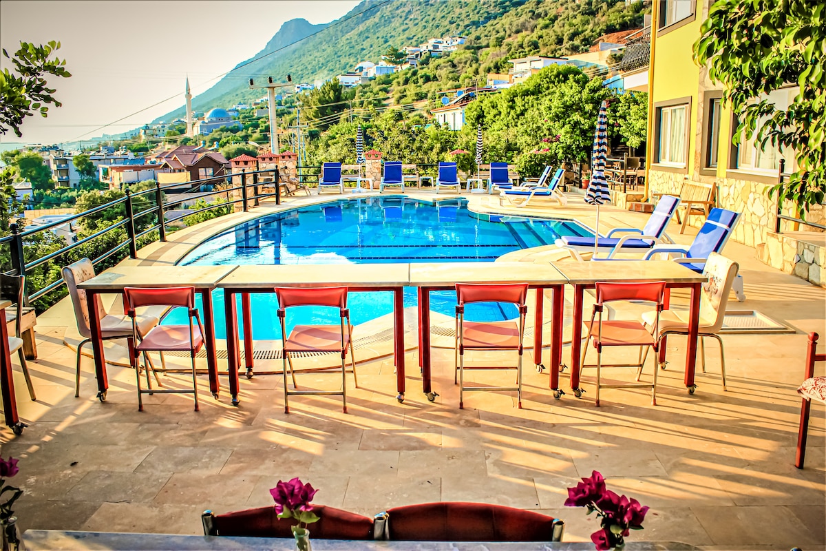 A spacious outdoor area features a swimming pool surrounded by sun loungers. A long table with multiple chairs is positioned nearby, complemented by vibrant flowers. Lush greenery and a hillside landscape create a scenic backdrop.