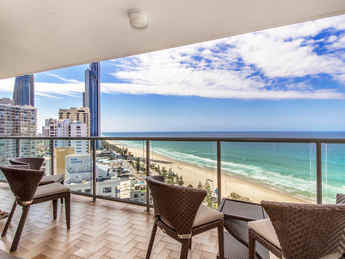 A spacious balcony offers unobstructed views of the beach and rolling waves. Comfortable seating is provided with wicker chairs positioned to enjoy the coastal scenery. The sky reflects a range of blues, enhancing the serene atmosphere of the beachfront location.