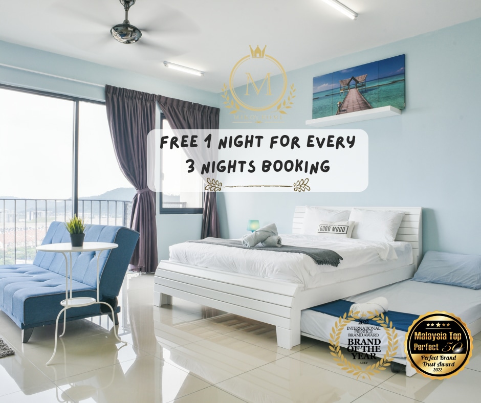 A bright studio space features a spacious bed with white linens and a decorative throw pillow. Natural light enters through a large window, and a comfortable seating area includes a blue chair and small table. Soft purple curtains frame the window, adding a touch of color.