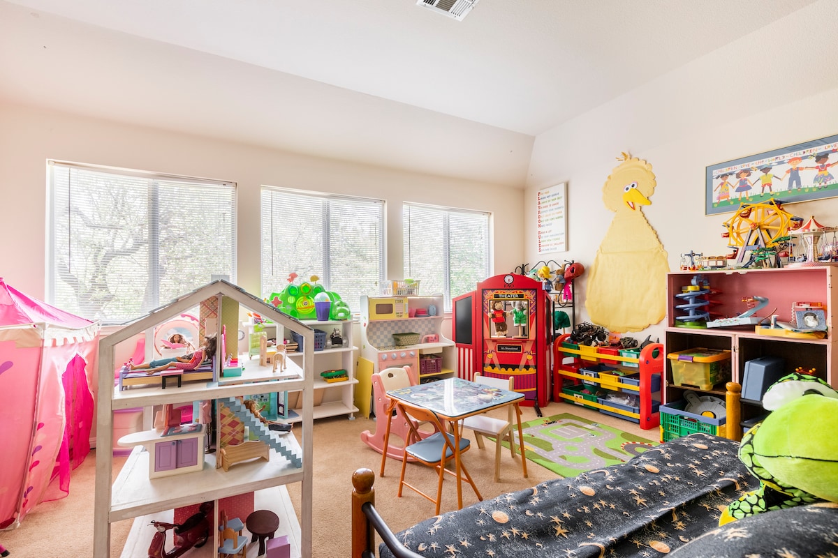 A vibrant children's room is filled with an assortment of colorful toys and play structures. Large windows allow natural light to illuminate the space featuring a playhouse, a small table, and various storage areas for toys. Bright decor, including a wall featuring a popular character, adds to the playful ambiance.