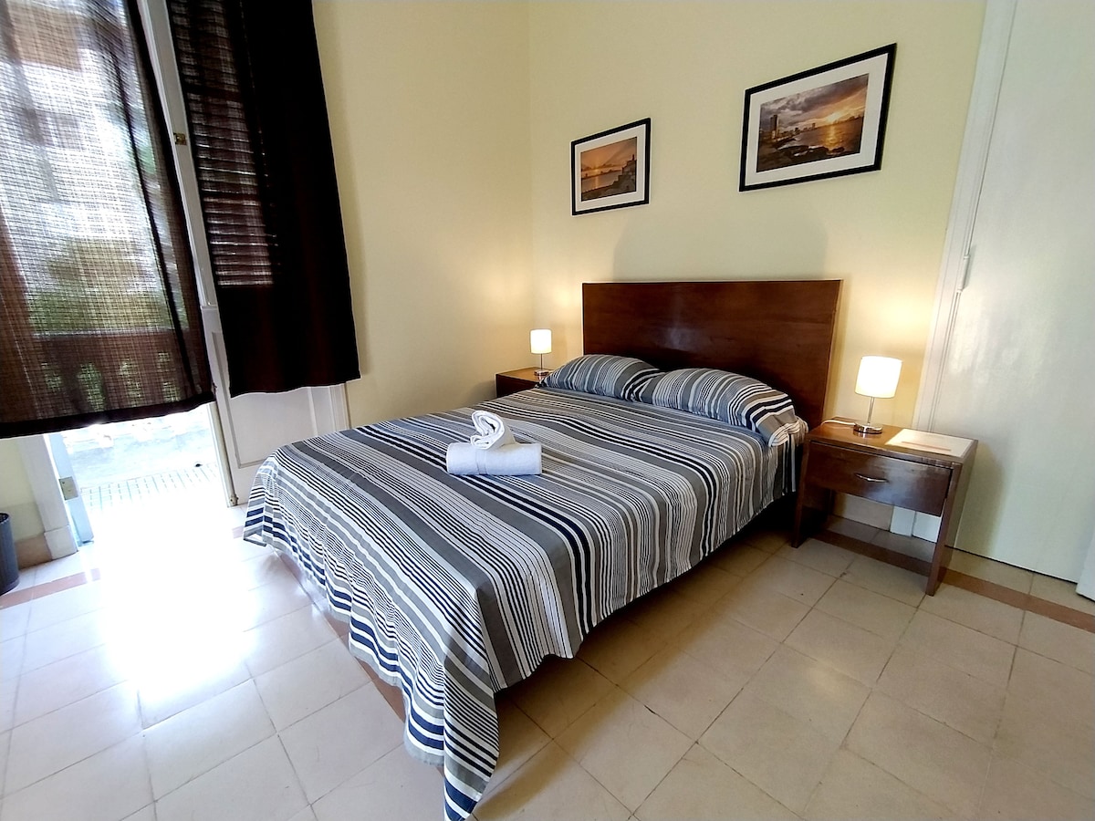 A comfortable bedroom is furnished with a wooden bed dressed in striped linens. Two bedside lamps offer soft lighting, and artwork adorns the walls. A sliding door leads to a balcony, providing access to fresh air and natural light. Tiled flooring complements the overall design.