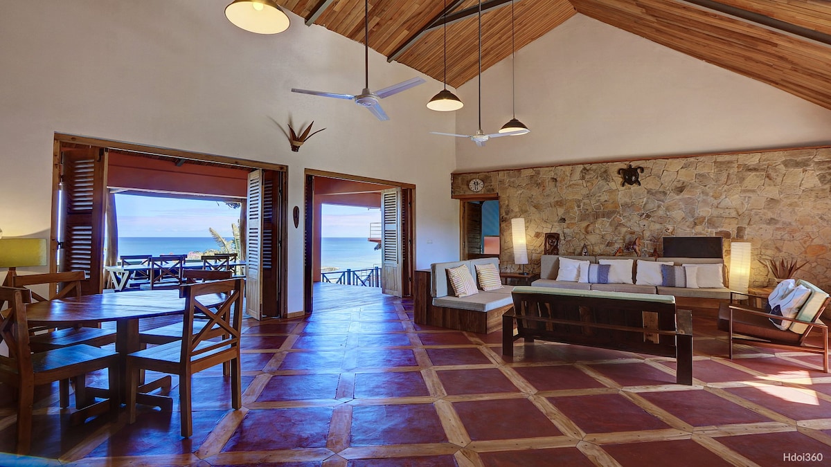 An open living area features a high wooden ceiling with decorative beams. Comfortable seating is arranged to face large windows offering a view of the ocean. A dining table is seen in the foreground, enhancing the spacious and inviting environment.
