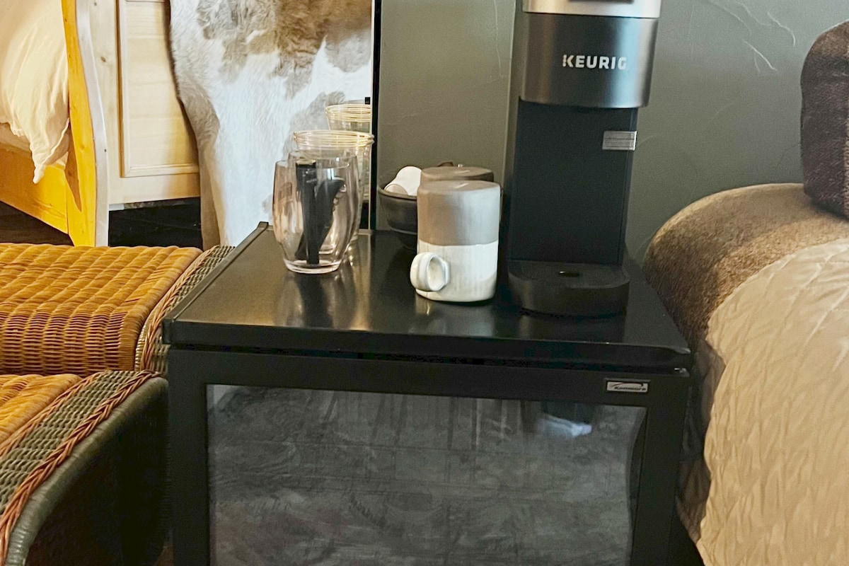 A small fridge sits beneath the Keurig coffee maker, which is placed on a black table. Several cups and glasses are neatly arranged next to the coffee maker, with a textured area rug visible in the background.