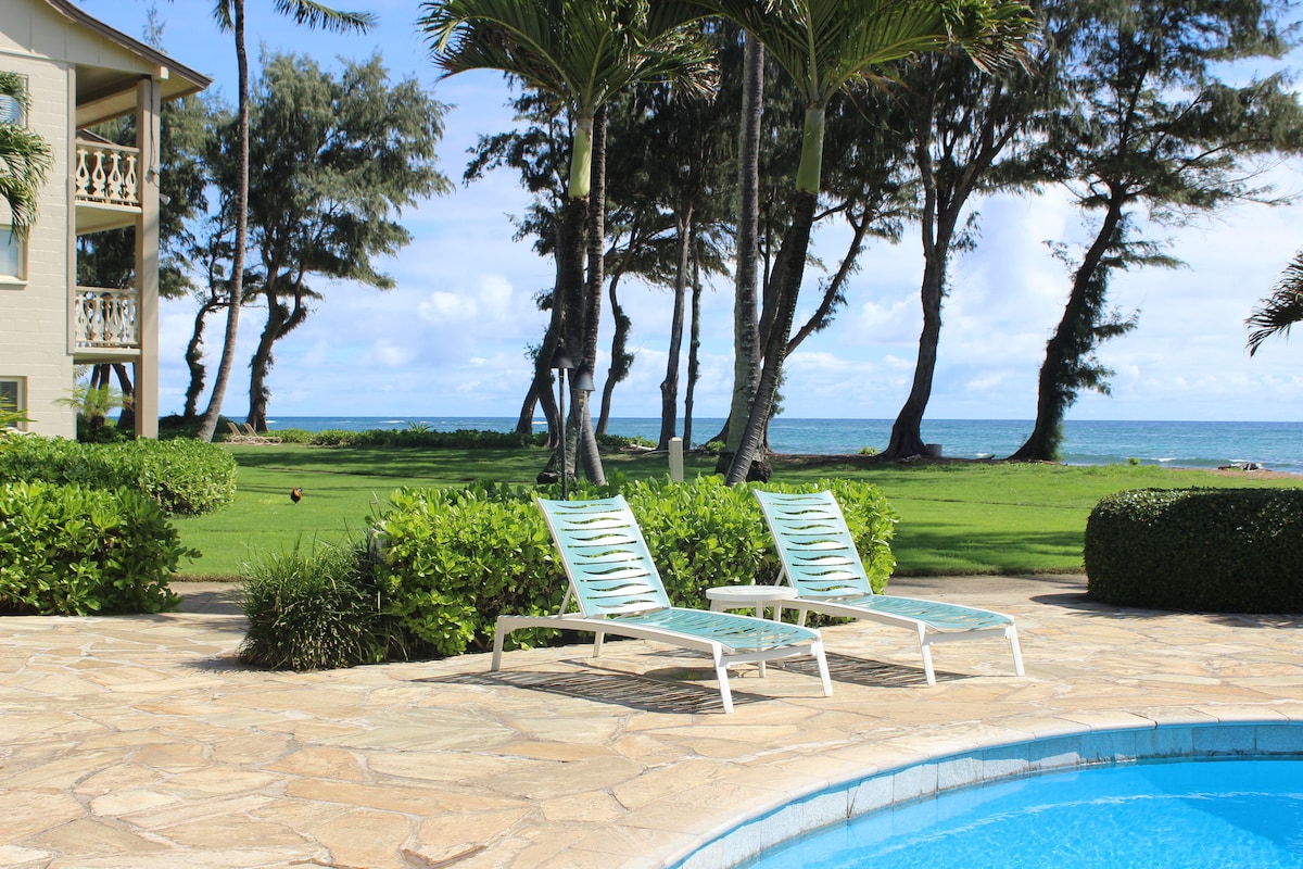 Two lounge chairs are positioned beside a clear blue pool, surrounded by a stone patio. Lush greenery and palm trees frame the scene, with ocean views visible through the trees. The inviting atmosphere features a serene landscape with a well-maintained lawn.