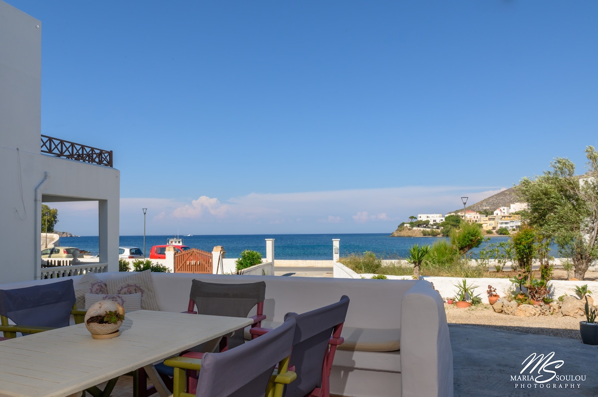 An outdoor seating area features a spacious table surrounded by comfortable chairs, offering views of the sea. Coastal greenery and a glimpse of the shoreline are visible in the background, creating a serene setting for relaxation.