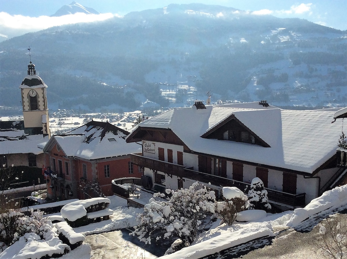 A snow-covered chalet building is visible in the foreground, complemented by a neighboring structure displaying a sign. Mountains rise majestically in the background, partially shrouded by clouds. The scene displays a serene winter landscape, accentuated by the quiet charm of the village.