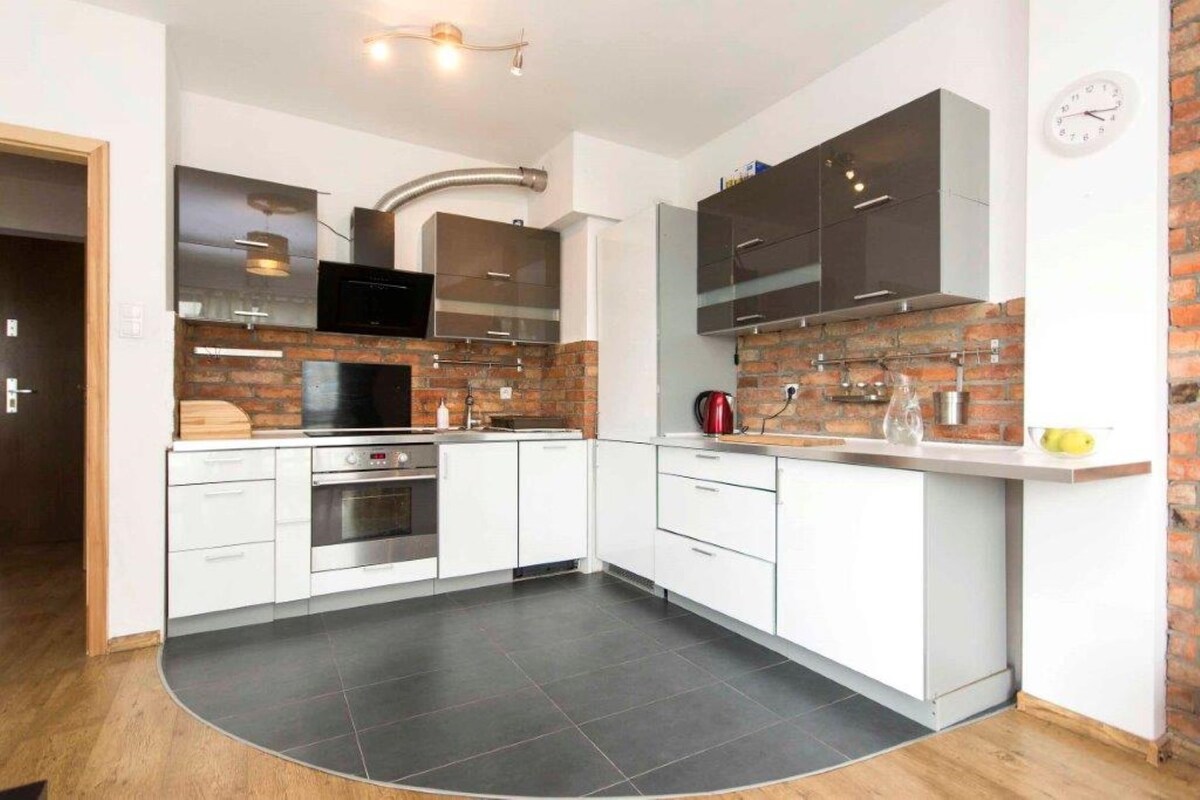 A modern kitchen is presented with a mix of sleek white and dark cabinets, complemented by exposed brick walls. Stainless steel appliances are visible, including an oven and a microwave. The countertop features a kettle and glassware, with a warm wooden floor enhancing the space.
