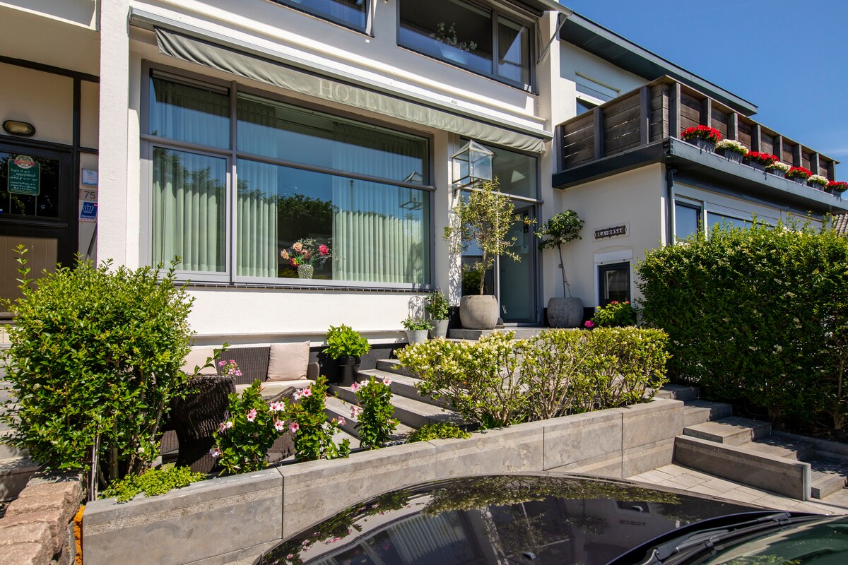 The hotel exterior features a clean, modern façade with large windows allowing natural light to filter through. Lush greenery and colorful flower arrangements enhance the entryway, which is framed by symmetrical planters. A spacious patio area is visible, offering comfortable seating options.