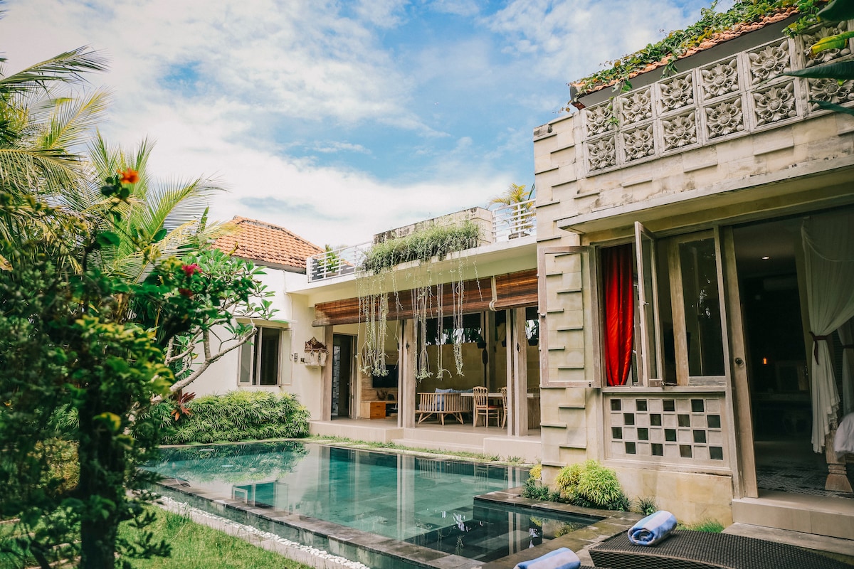 A tranquil villa is surrounded by lush greenery and features a private pool. The outdoor space creates a seamless connection with nature, while the building showcases an elegant design with expansive glass doors and natural light illuminating the interiors.