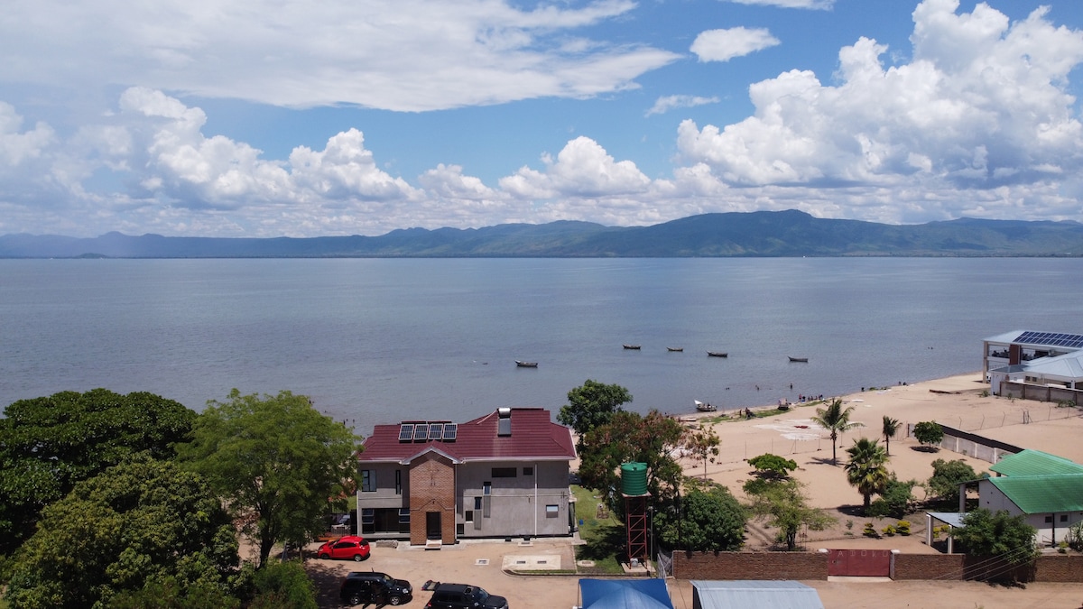 A panoramic view showcases the modern cottage alongside the beachfront, with expansive waters of Lake Malawi in the foreground. The serene landscape features clouds reflecting sunlight over the lake, while fishing boats and palm trees dot the sandy shore, creating a tranquil atmosphere.