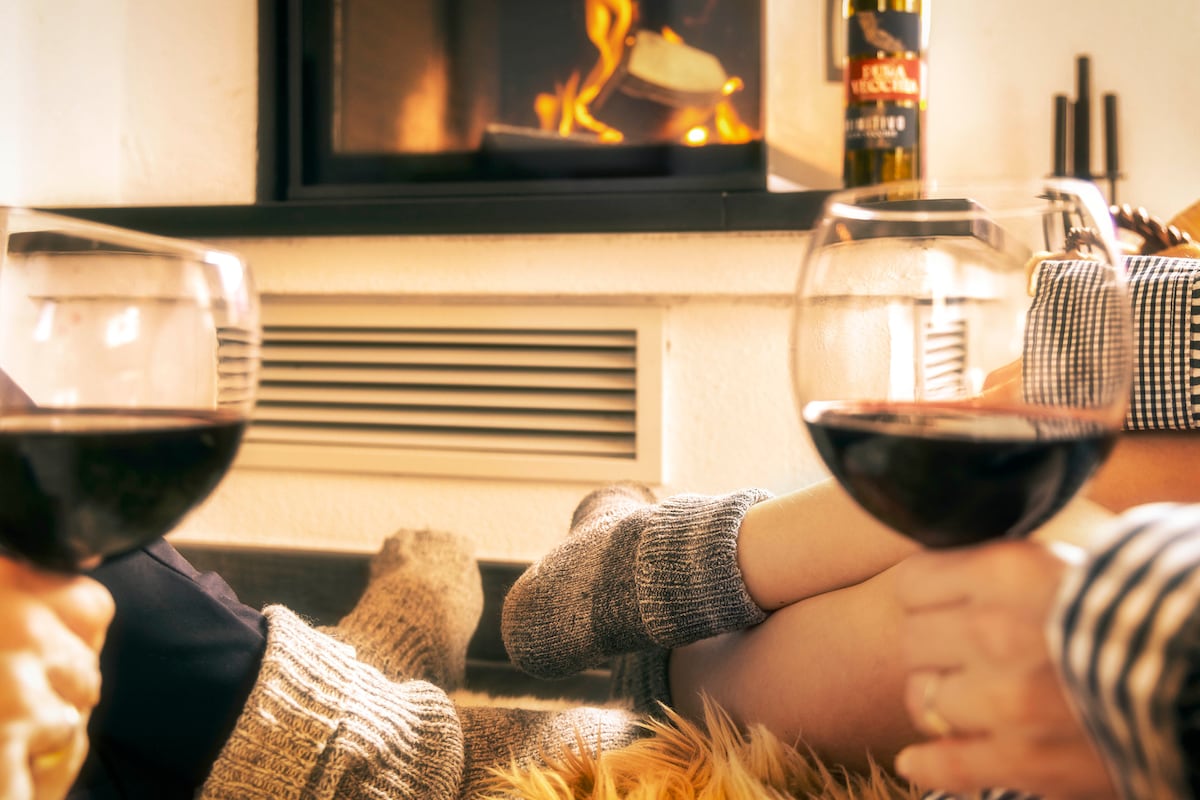 Two pairs of hands hold glasses of red wine, positioned near a cozy fireplace. A warm blanket is draped across the lower half of the image, complemented by soft socks. The flickering flames in the fireplace create a relaxing ambiance.