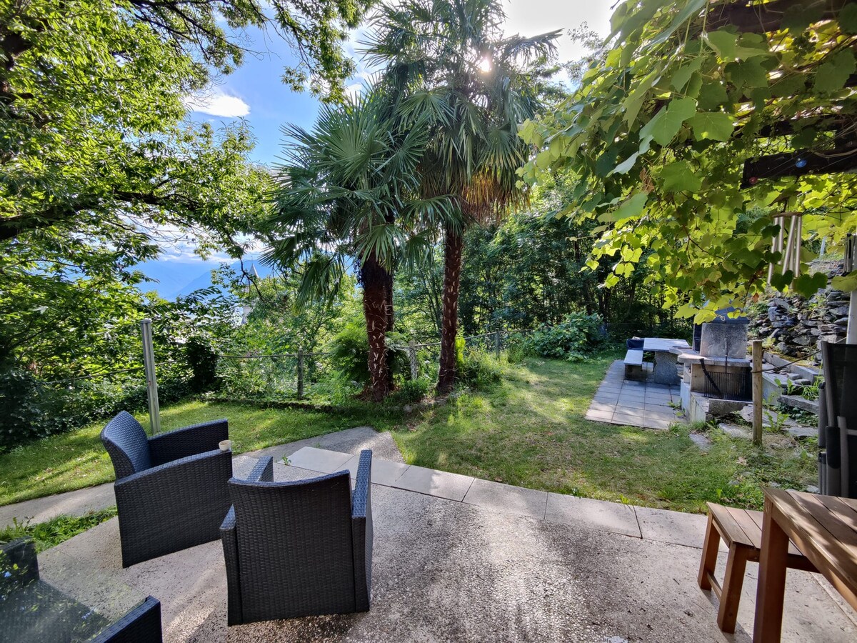 A private garden space is showcased, featuring two wicker chairs arranged around a small table. Lush greenery surrounds the area, with tall palm trees and other foliage visible. The stone path leads to a shaded spot, enhancing the tranquil outdoor setting.