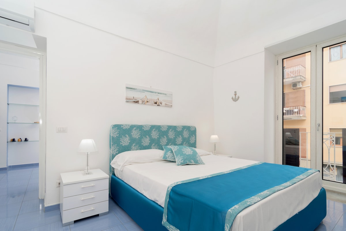 A spacious bedroom is furnished with a large bed featuring a teal and white design. Two bedside tables with lamps are placed beside the bed. Natural light enters through the glass doors leading to a small balcony. A nautical-themed decor piece is displayed on the wall.