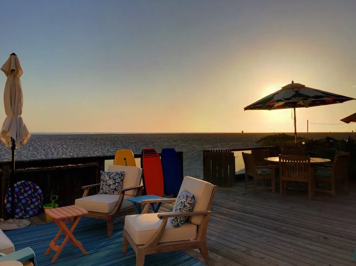 An oceanfront deck is featured, furnished with comfortable seating and a small table, providing a serene space for relaxation. Brightly colored boogie boards are positioned against a wall, and a large umbrella offers shade. The sunset casts a warm glow over the tranquil beach view.