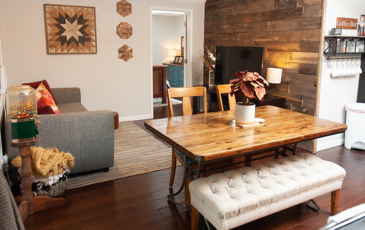 A dining area features a rustic wooden table surrounded by four chairs, complemented by a cushioned bench. A cozy couch is visible in the background. The wall showcases natural wood paneling and decorative accents, with a television mounted nearby, creating a stylish space for relaxation and dining.