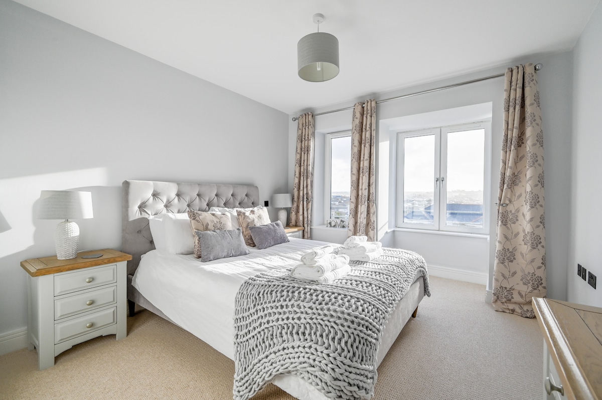 A spacious master bedroom features a king-size bed with a stylish gray headboard, accompanied by plush pillows and an inviting throw. Two large windows allow natural light to enter, complemented by soft curtains. A lamp stands on a bedside table, enhancing the room's tranquil ambiance.