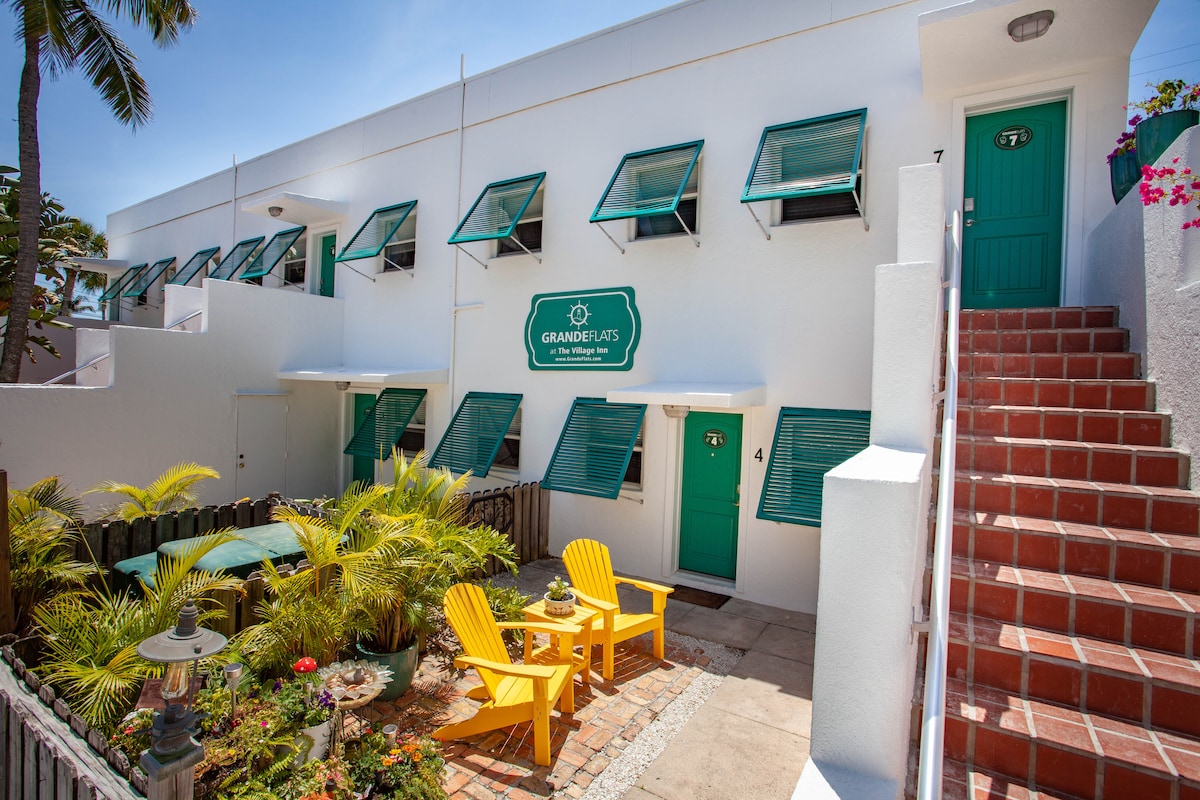 A bright exterior view of a two-story building is displayed, featuring green shutters and doors. Yellow adirondack chairs are positioned in a landscaped courtyard, alongside tropical plants. An inviting stone pathway leads to the entrance, complemented by clear blue skies overhead.
