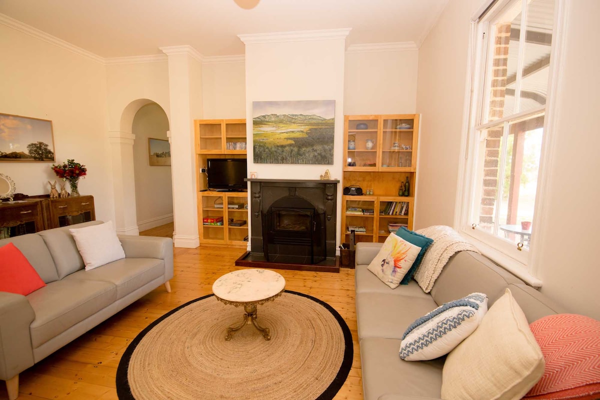 A comfortable living area features polished wooden floors and bright natural light. Two sofas are positioned around a central round rug, with decorative cushions adding color. A classic fireplace sits beneath a landscape painting, complemented by a wooden shelf holding books and a television.