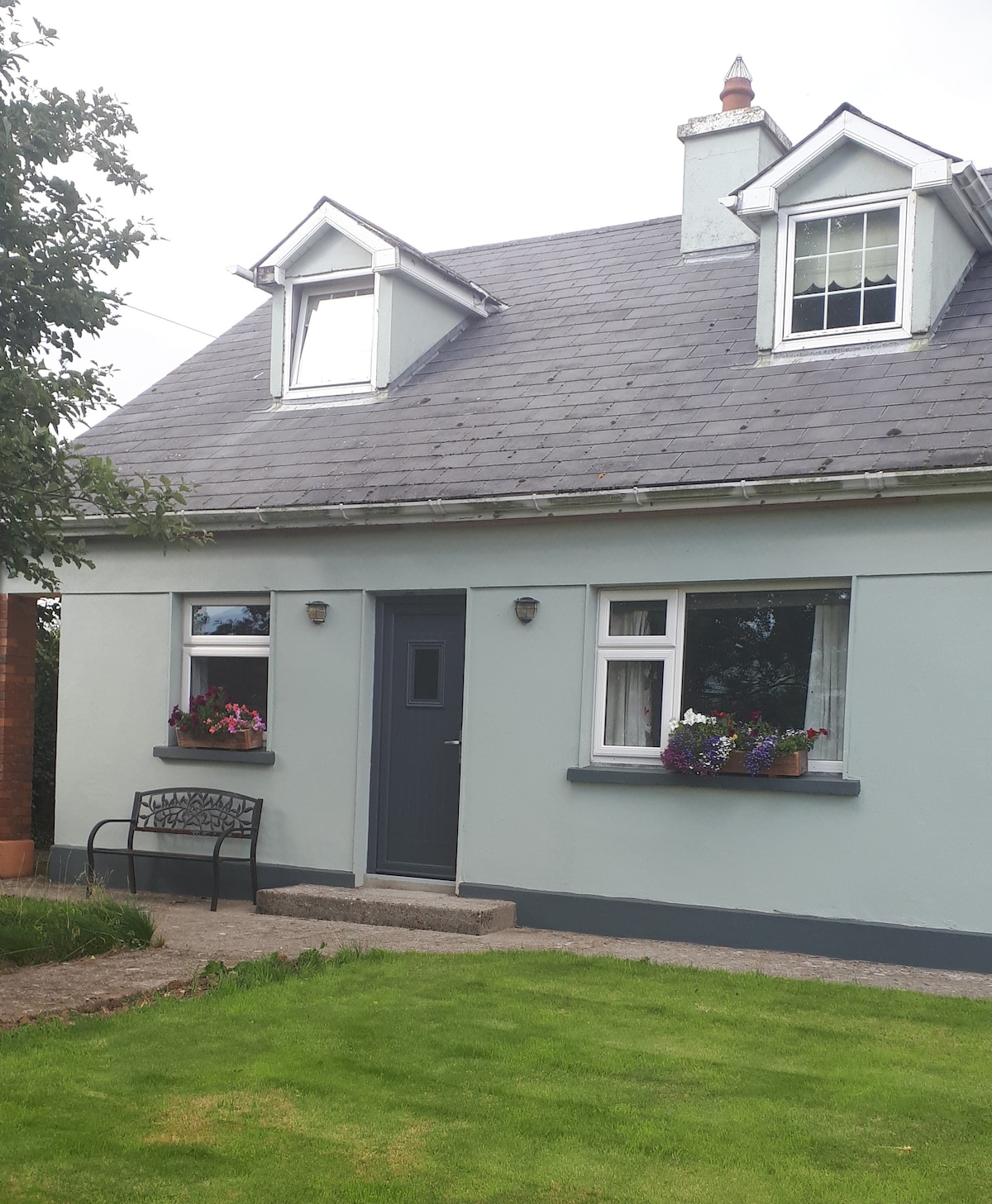 The exterior of a charming two-story home is presented, featuring a light blue facade with white trim. Two gabled windows add character, while flower boxes adorned with colorful blooms enhance the welcoming appeal. A paved path leads to the front door, flanked by a small grassy area.