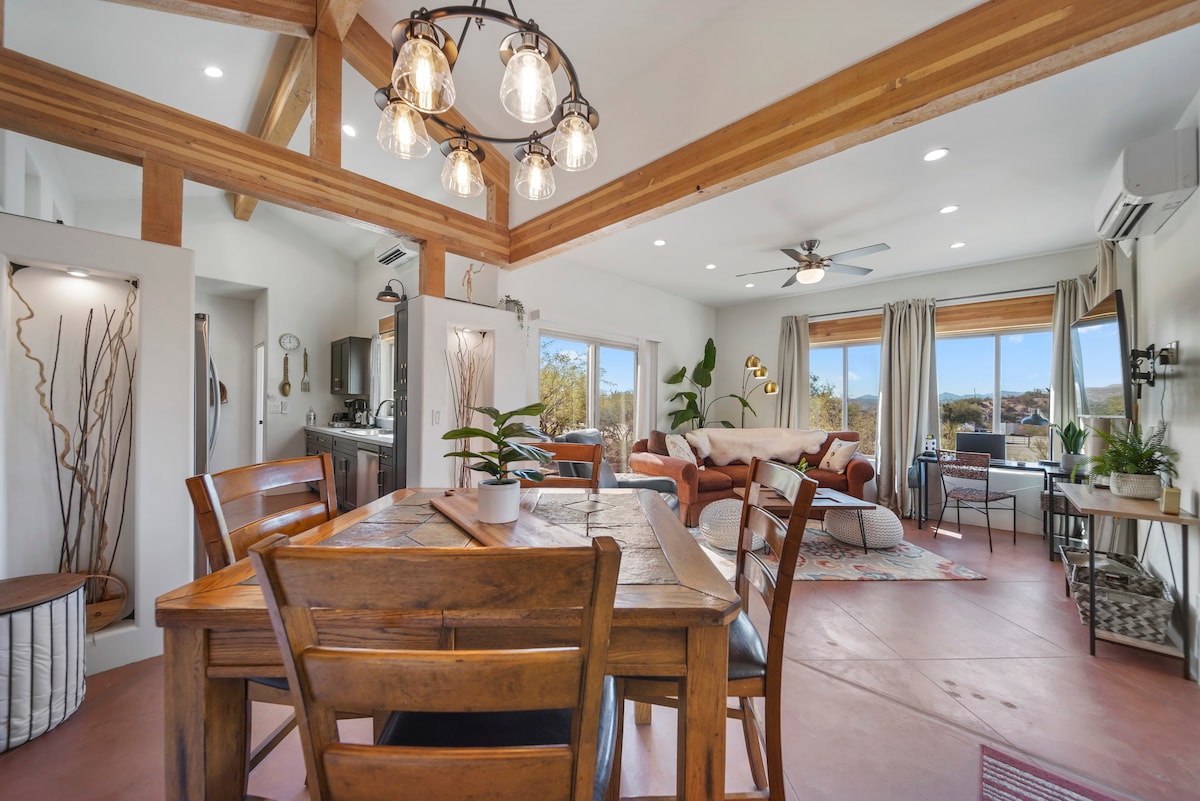 An open living space features a wooden dining table surrounded by chairs, with a cozy seating area in the background. Large windows offer natural light and views of the outdoor landscape. Exposed wooden beams and modern light fixtures add to the ambiance.