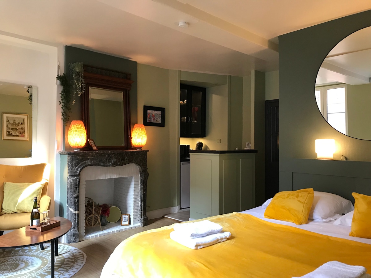 A spacious area features a bed with yellow bedding and soft pillows, complemented by a cozy chair in the corner. A decorative mirror reflects light, while a fireplace adds character. A kitchenette area is visible, showcasing modern cabinetry and various decorative elements.