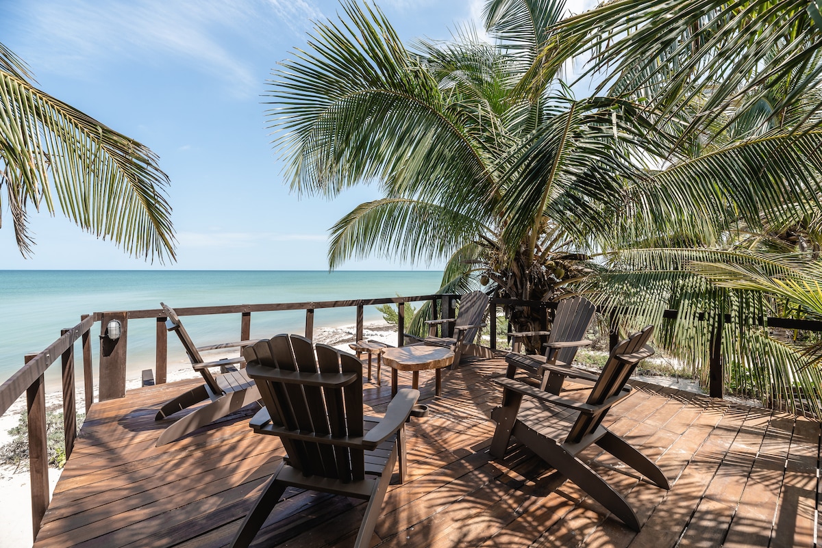 A wooden deck is positioned facing the beach and sea, featuring several adirondack chairs arranged around a circular table. Lush palm trees frame the area, providing shade. The calm waters and sandy shore are visible in the background, enhancing the natural setting.