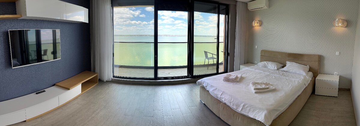 A spacious bedroom features a comfortable bed with fresh linen, complemented by a large window offering views of a serene waterfront. A balcony is visible, alongside a chair, creating a relaxing space to enjoy the natural surroundings. LCD TV and soft lighting enhance the inviting atmosphere.