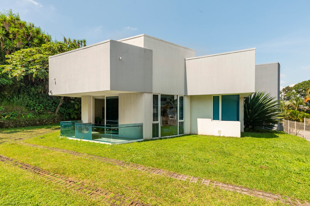 A modern two-story house is surrounded by lush greenery, showcasing large glass windows that invite natural light. A spacious patio area is visible, extending towards a well-maintained garden. The clean lines and minimalist design highlight a contemporary architectural style.