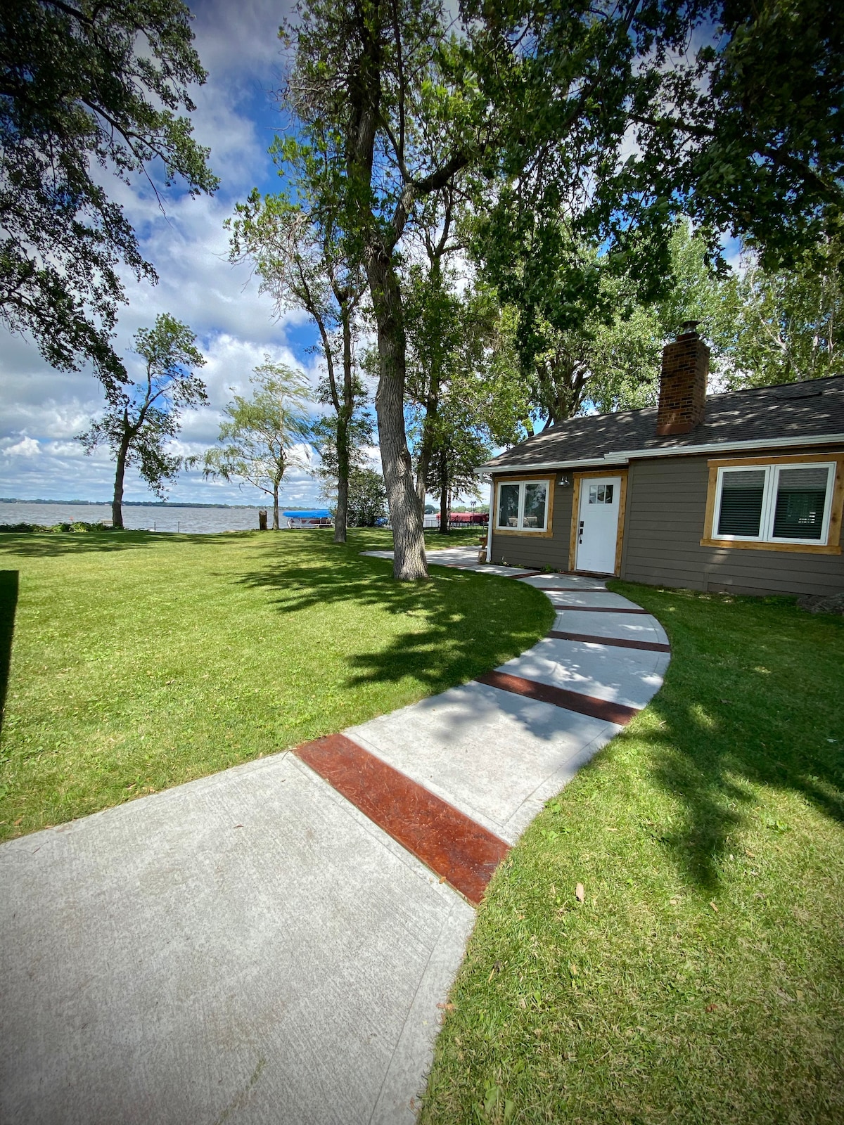 A path of concrete and wood leads from the cabin to the lakeside, framed by lush green grass and mature trees. The cabin features windows and a welcoming entrance, providing clear views of Lake Kampeska in the distance under a bright blue sky.