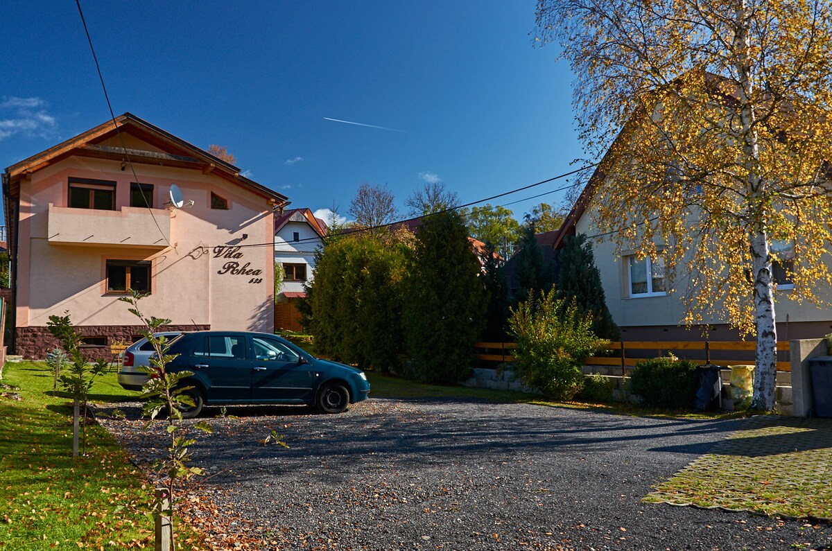 A welcoming building with a light pink facade and greenery surrounds it. A spacious parking area is available, featuring a parked car. The setting includes well-maintained hedges and a clear blue sky above, contributing to a serene environment.