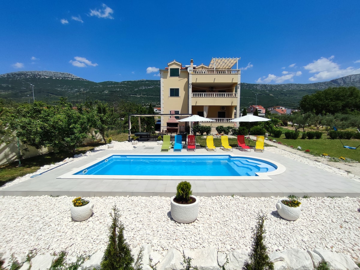 A spacious outdoor area features a swimming pool surrounded by colorful lounge chairs. The building, with multiple floors, is positioned in the background, showcasing a terrace. Lush greenery and a well-maintained garden enhance the setting, with clear blue skies above.