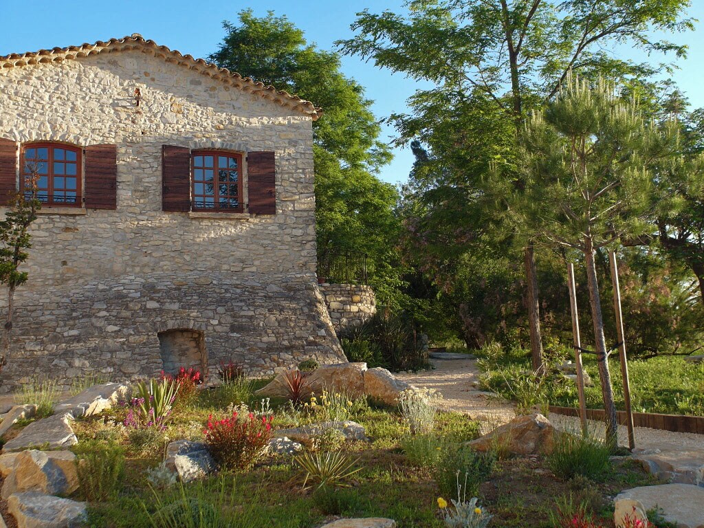 A charming stone house is framed by lush greenery. Two wooden windows with shutters are visible, complementing the natural stone facade. Flowering plants and shrubs are arranged in a landscaped area, enhancing the outdoor space beside a winding path.