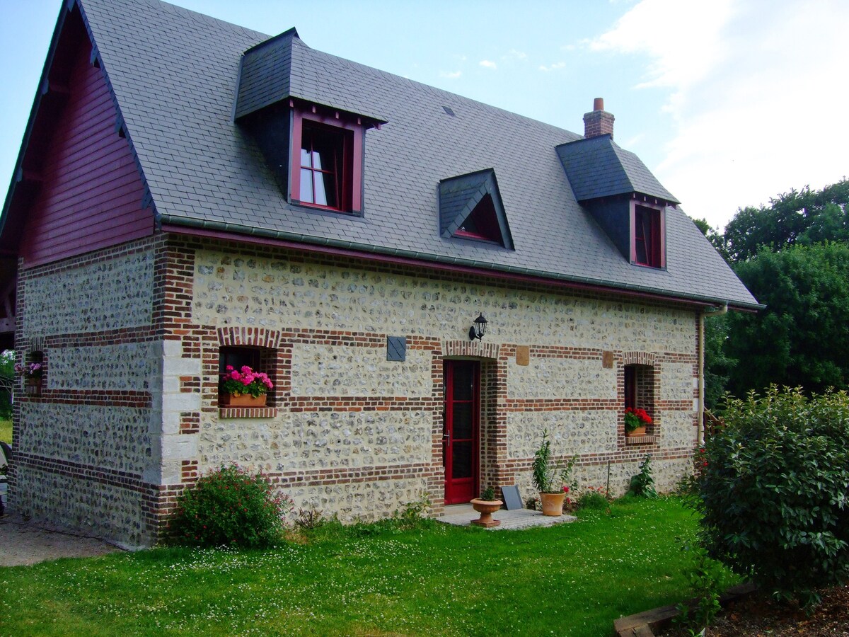 The charming stone exterior of the cottage is complemented by red window frames and flower boxes, showcasing vibrant plants. A manicured lawn surrounds the structure, adding to the serene environment. The sloped roof and unique window shapes enhance the property’s rustic character.